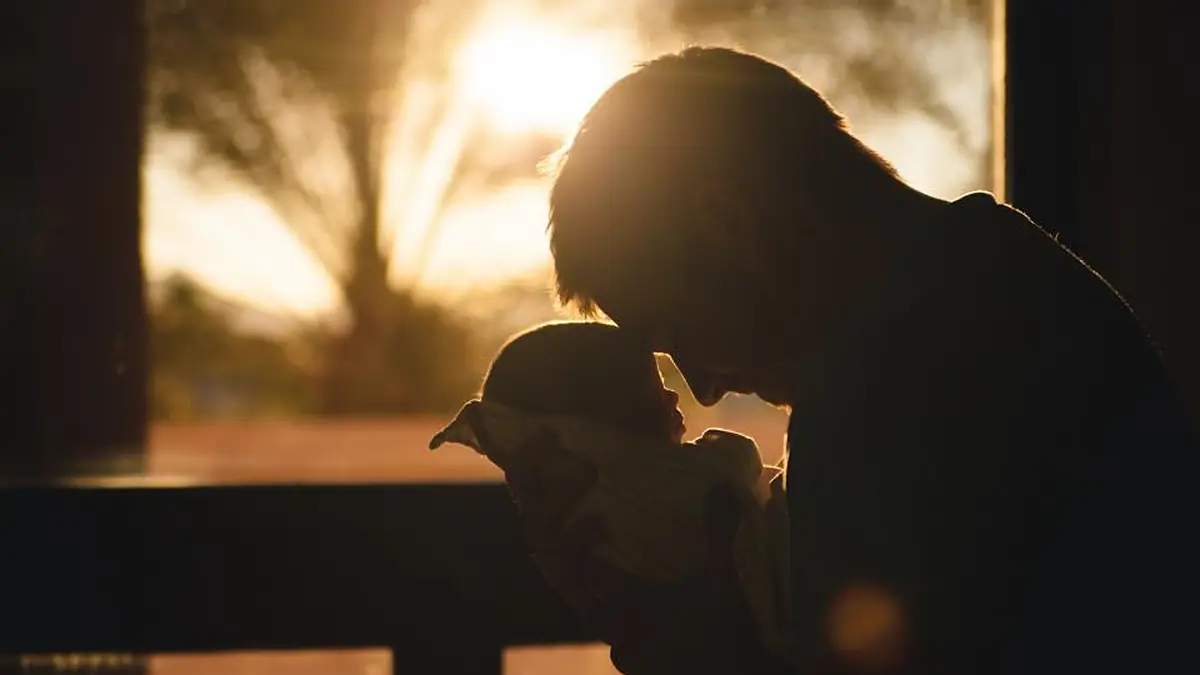 Silhueta de adulto com bebê no colo diante de uma janela ao pôr do sol
